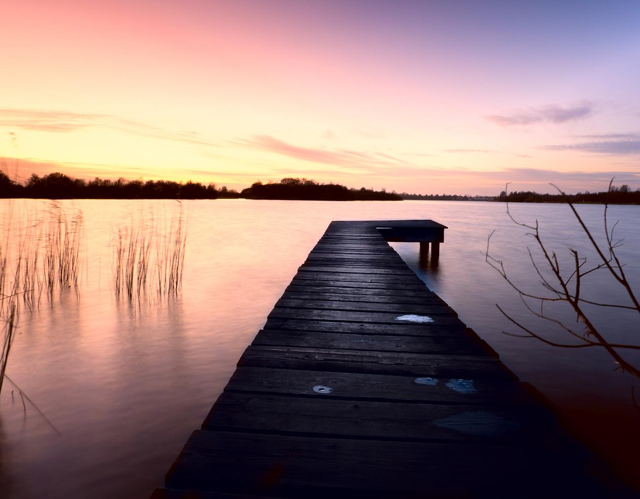 Holzsteg führt über ruhiges Wasser, umgeben von Gräsern und einem farbenprächtigen Sonnenuntergang.